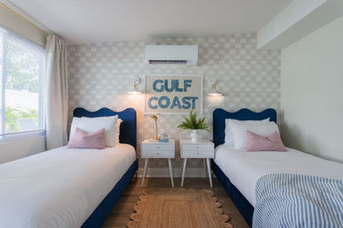 Bright Gulf Coast–themed twin bedroom with navy velvet headboards, crisp white bedding and pink accent pillows, matching white nightstands with brass wall lamps and a potted plant, framed “GULF COAST” wall art beneath a wall-mounted AC, window with curtains and a woven jute rug.