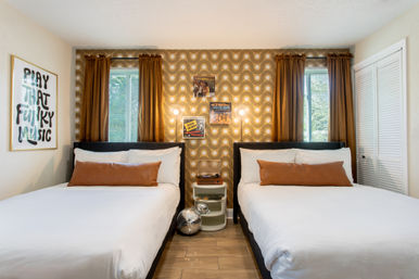 Retro-inspired guest room with two beds, white linens and brown bolster pillows, wavy 1970s wallpaper, record player on a nightstand, framed 'Play That Funky Music' art, disco balls and mustard curtains.