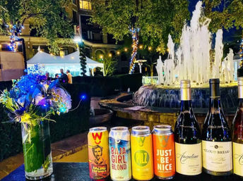 Nighttime hotel courtyard scene with illuminated fountain and string lights, tent in background; vase of LED-lit flowers and a lineup of colorful craft beer cans and wine bottles on a table.