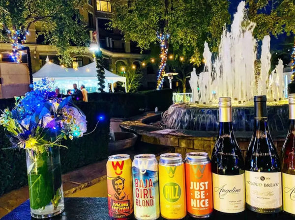 Nighttime hotel courtyard scene with illuminated fountain and string lights, tent in background; vase of LED-lit flowers and a lineup of colorful craft beer cans and wine bottles on a table.