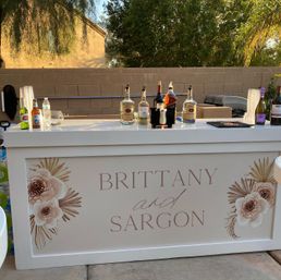 White customized outdoor wedding bar on a backyard patio with floral panels, liquor bottles, wine and stacked plastic cups