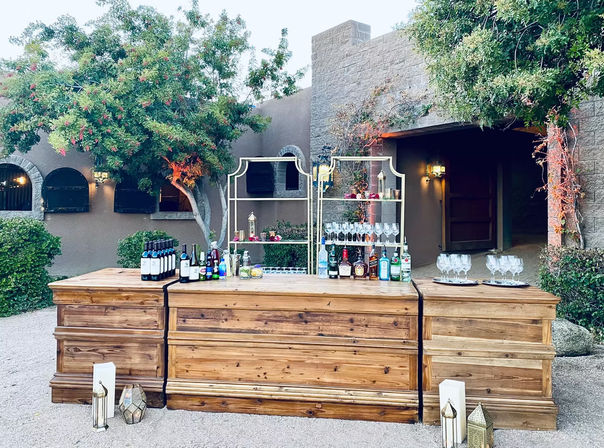 Rustic outdoor wooden bar in a Southwestern courtyard with stacked wood counters, wine and liquor bottles, glassware on decorative shelves, lanterns and leafy trees framing a stylish cocktail station.