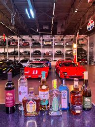 Liquor bottles on a bar counter with bright red sports cars in the foreground and stacked vintage and modern cars on multi-level racks inside a neon-lit warehouse-style car collection.