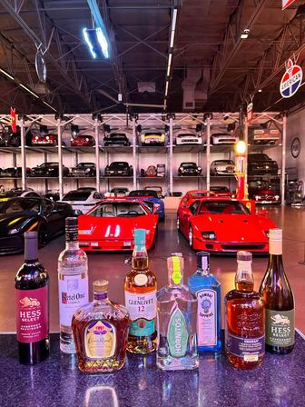 Liquor bottles on a bar counter with bright red sports cars in the foreground and stacked vintage and modern cars on multi-level racks inside a neon-lit warehouse-style car collection.