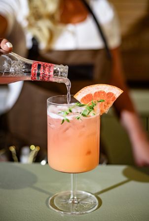 Close-up of a bartender pouring sparkling water into a zesty pink grapefruit cocktail, garnished with a grapefruit wedge and fresh herb in a stemmed glass on a bar