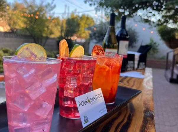 Three colorful iced cocktails with lime and citrus garnishes on a black tray atop a wooden patio table, wine bottles and twinkling string lights in a backyard evening.