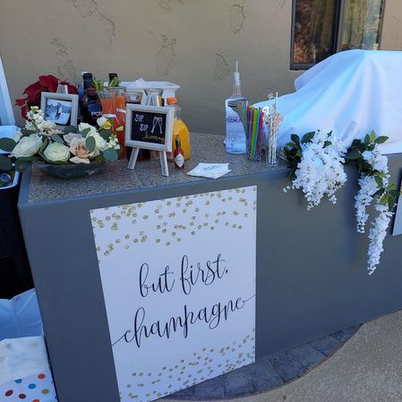 Outdoor patio champagne and mimosa bar setup with sign reading "but first, champagne", white floral arrangements, carafes of juices, cocktail mixers and colorful straws on a stone countertop