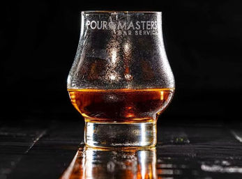 Close-up of a branded whiskey tasting glass with rich amber liquid, beaded condensation and a glowing reflection on dark wet wood.