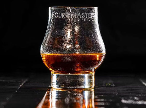 Close-up of a branded whiskey tasting glass with rich amber liquid, beaded condensation and a glowing reflection on dark wet wood.