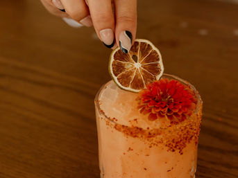 Hand placing a dried lime slice on a pink craft cocktail with crushed ice, chili-spiced rim and red edible flower garnish on a wooden table.