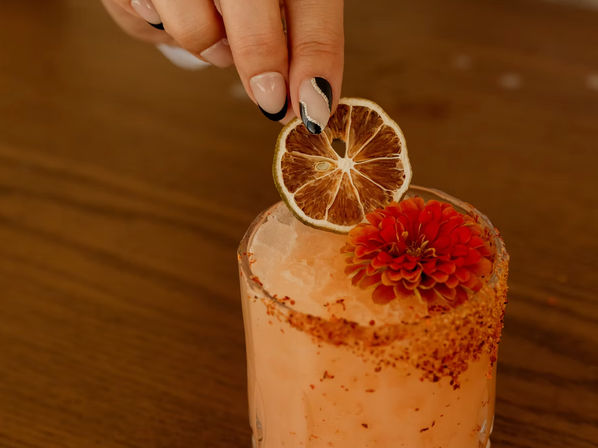 Hand placing a dried lime slice on a pink craft cocktail with crushed ice, chili-spiced rim and red edible flower garnish on a wooden table.