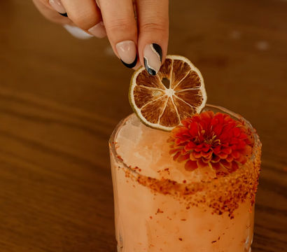 Hand placing a dried lime slice on a pink craft cocktail with crushed ice, chili-spiced rim and red edible flower garnish on a wooden table.