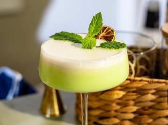 Frothy pale-green craft cocktail in a coupe glass, garnished with fresh mint leaves and a dried lime wheel, styled on a bar surface with wicker basket and cocktail tools in the background.
