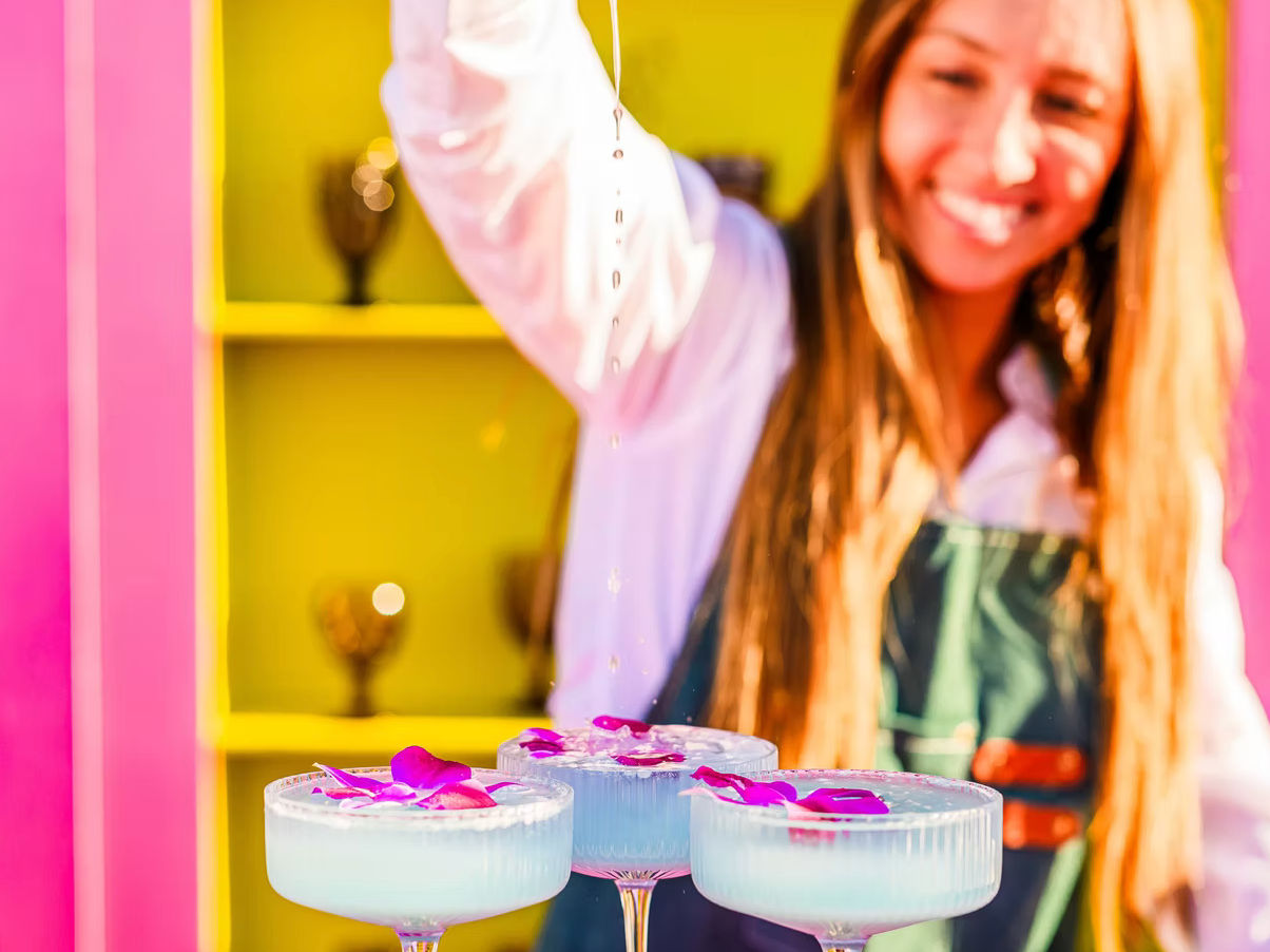Three pale blue craft cocktails in coupe glasses topped with purple orchid petals while a smiling bartender drizzles liquid over them against a vibrant pink and yellow bar backdrop