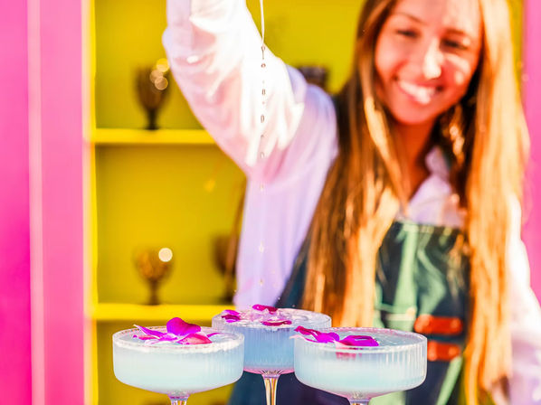 Three pale blue craft cocktails in coupe glasses topped with purple orchid petals while a smiling bartender drizzles liquid over them against a vibrant pink and yellow bar backdrop