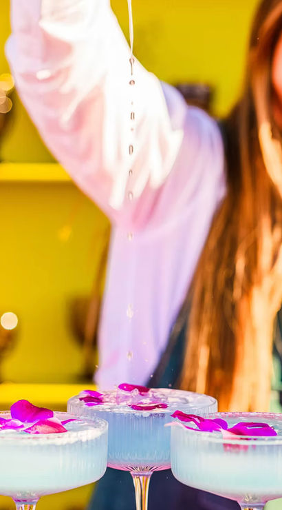 Three pale blue craft cocktails in coupe glasses topped with purple orchid petals while a smiling bartender drizzles liquid over them against a vibrant pink and yellow bar backdrop