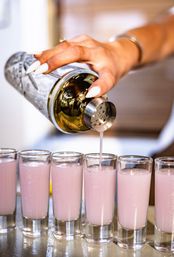 Close-up of a hand with white nails pouring pink creamy cocktail shots from a metal shaker into a row of glass shot glasses