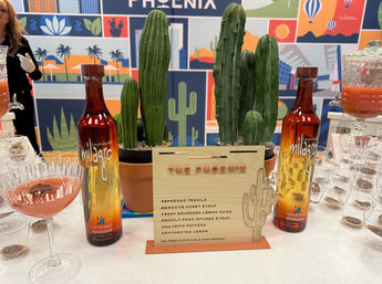 Desert-themed Phoenix cocktail station with two amber-to-gold spirit bottles flanking potted columnar cacti, a wooden cocktail menu, a pink coupe cocktail glass, and a colorful desert mural backdrop.