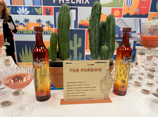 Desert-themed Phoenix cocktail station with two amber-to-gold spirit bottles flanking potted columnar cacti, a wooden cocktail menu, a pink coupe cocktail glass, and a colorful desert mural backdrop.