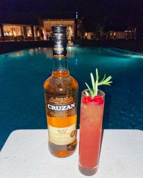 Poolside at night — a bottle of aged rum and a tall pink cocktail garnished with cherries and rosemary on a table beside a glowing resort pool.
