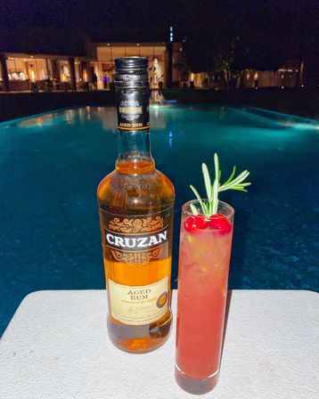 Poolside at night — a bottle of aged rum and a tall pink cocktail garnished with cherries and rosemary on a table beside a glowing resort pool.