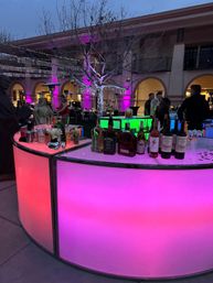 Evening outdoor courtyard party with a curved LED bar glowing pink and green, bottles on the counter, a leafless tree wrapped in string lights and silver tinsel, purple uplighting on arched building facades and people mingling.