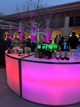 Evening outdoor courtyard party with a curved LED bar glowing pink and green, bottles on the counter, a leafless tree wrapped in string lights and silver tinsel, purple uplighting on arched building facades and people mingling.