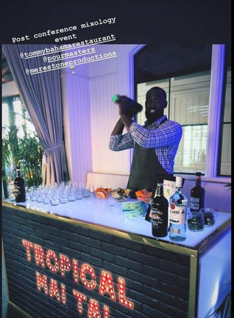 Indoor post-conference mixology scene: bartender shaking a cocktail behind a lit “Tropical Mai Tai” bar station lined with rum bottles, glassware and citrus garnishes — lively cocktail service at an event.