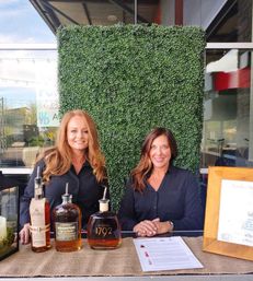 Two smiling women at an outdoor patio bourbon tasting table with three whiskey bottles (Basil Hayden, Redemption, 1792), tasting notes and a green hedge backdrop — casual restaurant patio event.