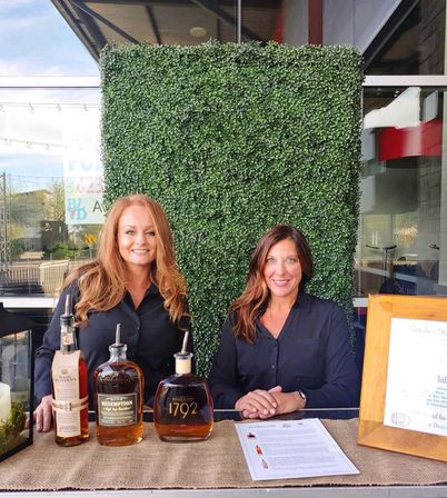 Two smiling women at an outdoor patio bourbon tasting table with three whiskey bottles (Basil Hayden, Redemption, 1792), tasting notes and a green hedge backdrop — casual restaurant patio event.