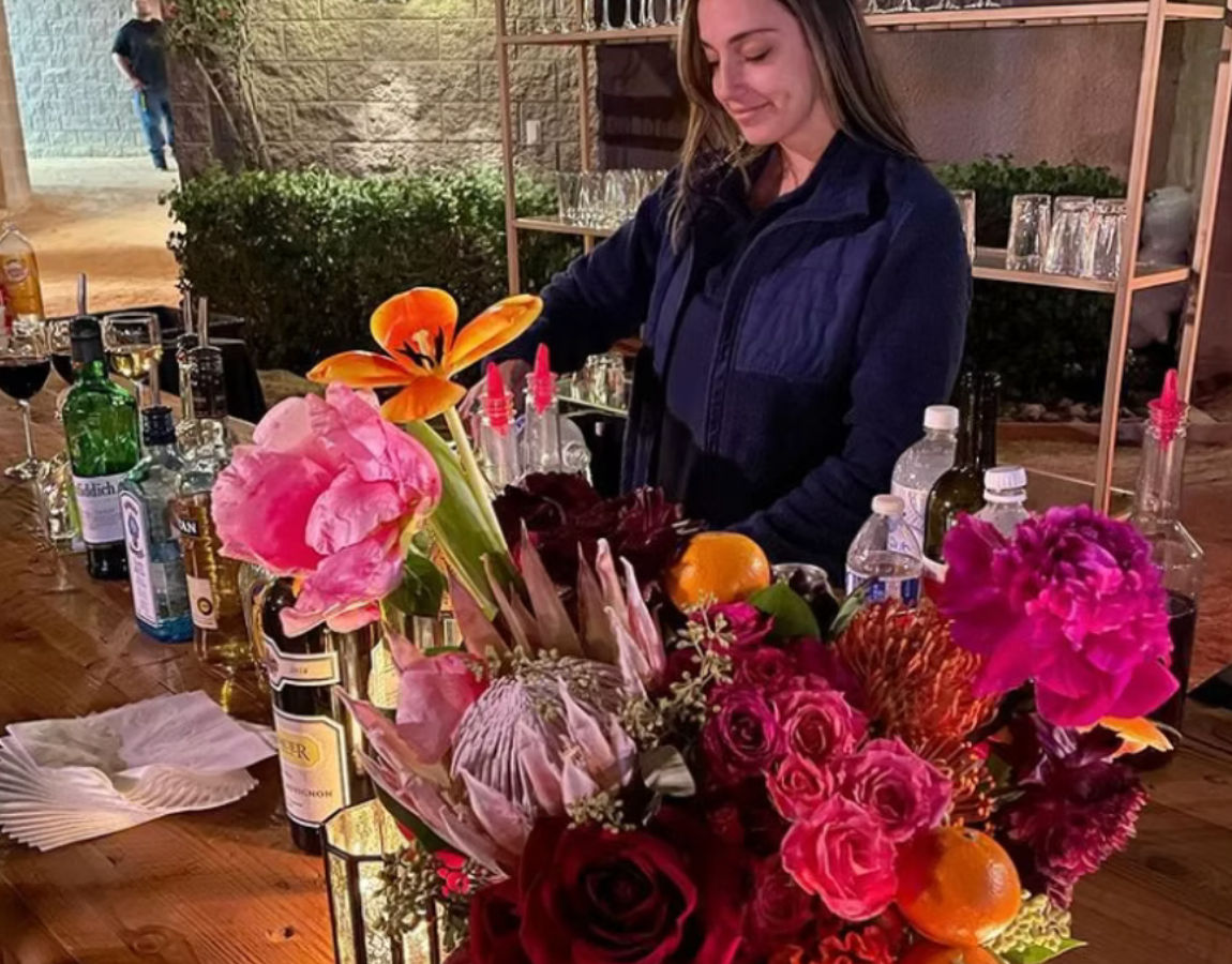 Outdoor patio bar scene with a person arranging a vibrant floral centerpiece of pink peonies, orange lilies and red roses on a wooden counter surrounded by bottles, glassware and citrus