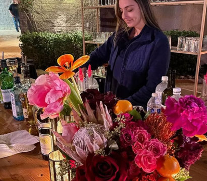 Outdoor patio bar scene with a person arranging a vibrant floral centerpiece of pink peonies, orange lilies and red roses on a wooden counter surrounded by bottles, glassware and citrus