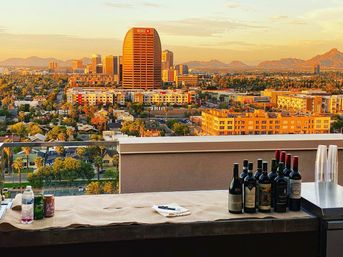 Rooftop golden-hour city skyline with distant mountains, foreground table set with wine bottles, cups and canned drinks — urban sunset view.