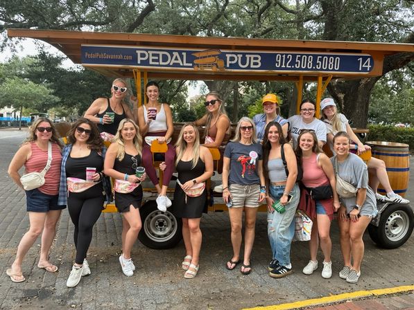 Group of women smiling and posing around a yellow pedal-powered party bike under live oak trees on a street in Savannah, GA, holding cups during a daytime celebration.