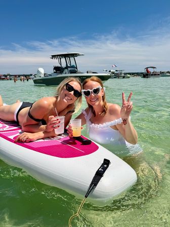 Two friends enjoying a summer beach day on a pink paddleboard in shallow clear coastal water, wearing sunglasses and swimsuits, holding cold drinks and smiling while one flashes a peace sign, with anchored boats and a sunny blue sky in the background.