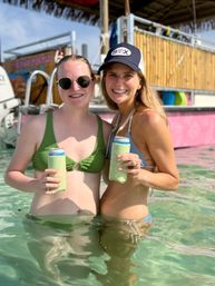 Two smiling women in bikinis standing waist-deep in clear tropical water holding canned drinks in green koozies, one wearing round sunglasses and the other a baseball cap, tiki-style beach bar and boat in the sunny background.