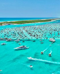 Aerial view of hundreds of boats and yachts anchored in crystal-clear turquoise water around a sandy island sandbar, a crowded dayboat party scene with speedboats carving white wakes under a bright blue sky