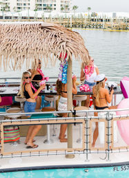 Group of women enjoying a tiki-roofed pontoon boat party in a Florida marina, wearing leis and sunglasses and holding pink cups with docks and waterfront condos in the background.