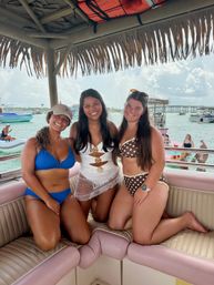 Three smiling friends in bikinis posing under a thatched tiki roof on a party boat with turquoise water, nearby boats and a distant bridge on a sunny tropical day