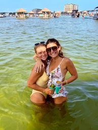 Two friends in swimsuits smiling and hugging waist-deep in green bay water, holding canned drinks with tiki huts and waterfront buildings in the sunny background.