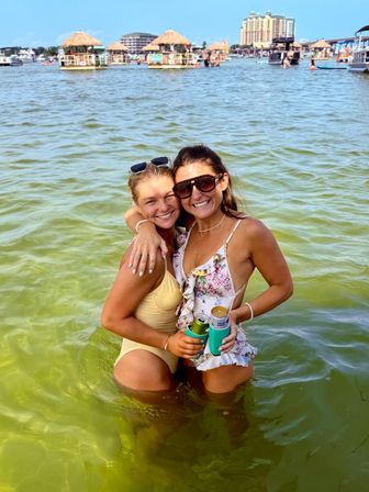 Two friends in swimsuits smiling and hugging waist-deep in green bay water, holding canned drinks with tiki huts and waterfront buildings in the sunny background.