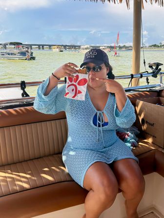 Person in blue crochet cover-up and 'Best Dad Ever' cap eating a sandwich from a red-logo bag on a boat, pointing to their mouth with a bridge and other boats on a sunny coastal bay in the background.