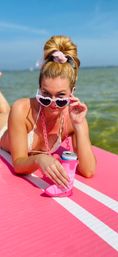 Person lounging on a pink paddleboard in shallow coastal water on a sunny summer day, peeking over heart-shaped pink sunglasses and holding a canned drink in a pink boot-shaped koozie.
