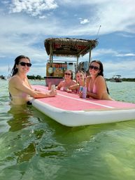 Four friends in bikinis relaxing on a pink paddleboard in shallow green water by a floating tiki bar, holding canned drinks under a sunny blue sky — summer beach day.