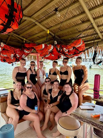 Group of women in swimsuits relaxing on a shaded party boat beneath hanging orange life jackets, docked in a sunny tropical bay with boats and a pier visible in the background.