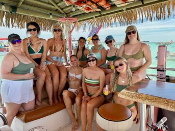 Smiling group of women in swimsuits at a thatched tiki boat bar holding orange cocktails, turquoise tropical water and boats in the background.
