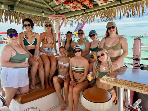 Smiling group of women in swimsuits at a thatched tiki boat bar holding orange cocktails, turquoise tropical water and boats in the background.