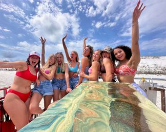 Eight friends in colorful bikinis and denim shorts cheer and raise their arms on a sunny white‑sand beach with blue skies and sand dunes