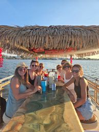 Seven friends smiling around a marble-pattern bar under a thatched tiki roof on a boat tiki-bar at sunset, drinks on the counter and coastal homes across a calm bay.