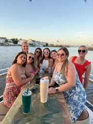 Group of eight friends in summer dresses and sunglasses smiling around a table with drinks on a boat at sunset, calm harbor and waterfront houses in the background — coastal, casual outing.